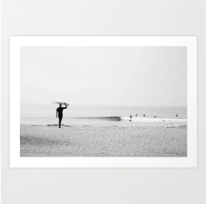 White  framed black and white photo of a person holding an surfboard on a beach with a minimalistic frame.